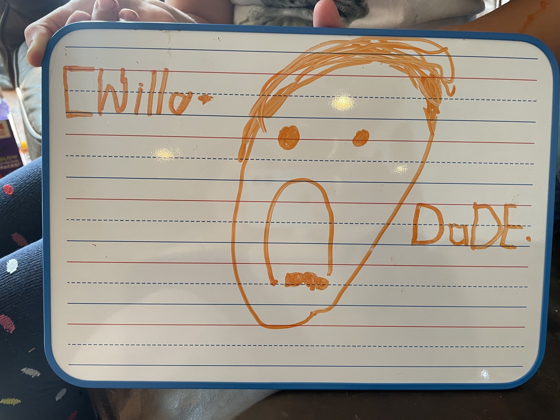 A lined handheld whiteboard filled with a child’s orange drawing and early writing.