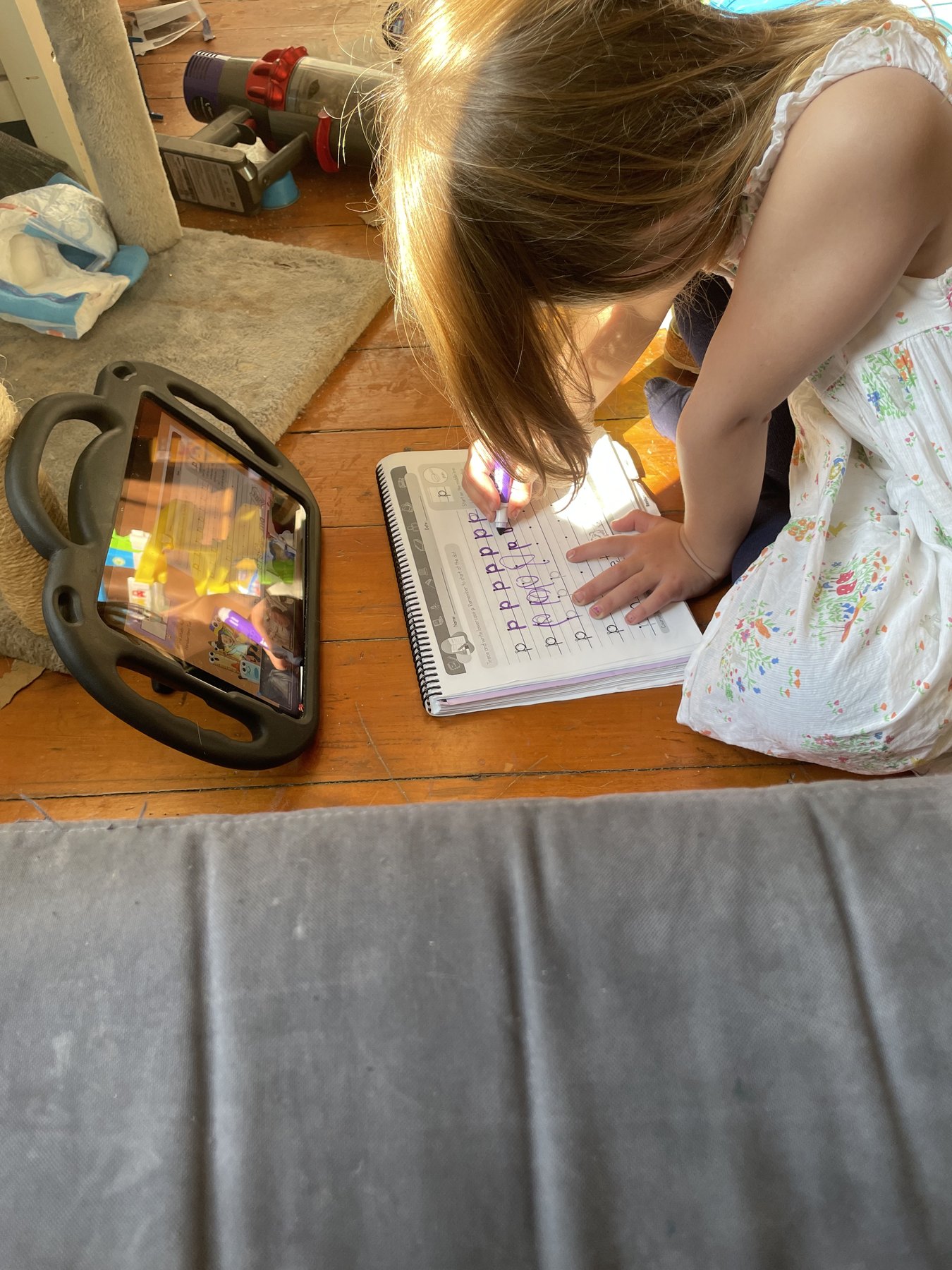 A young child watches a lesson on a protected tablet while completing handwriting work on the floor.